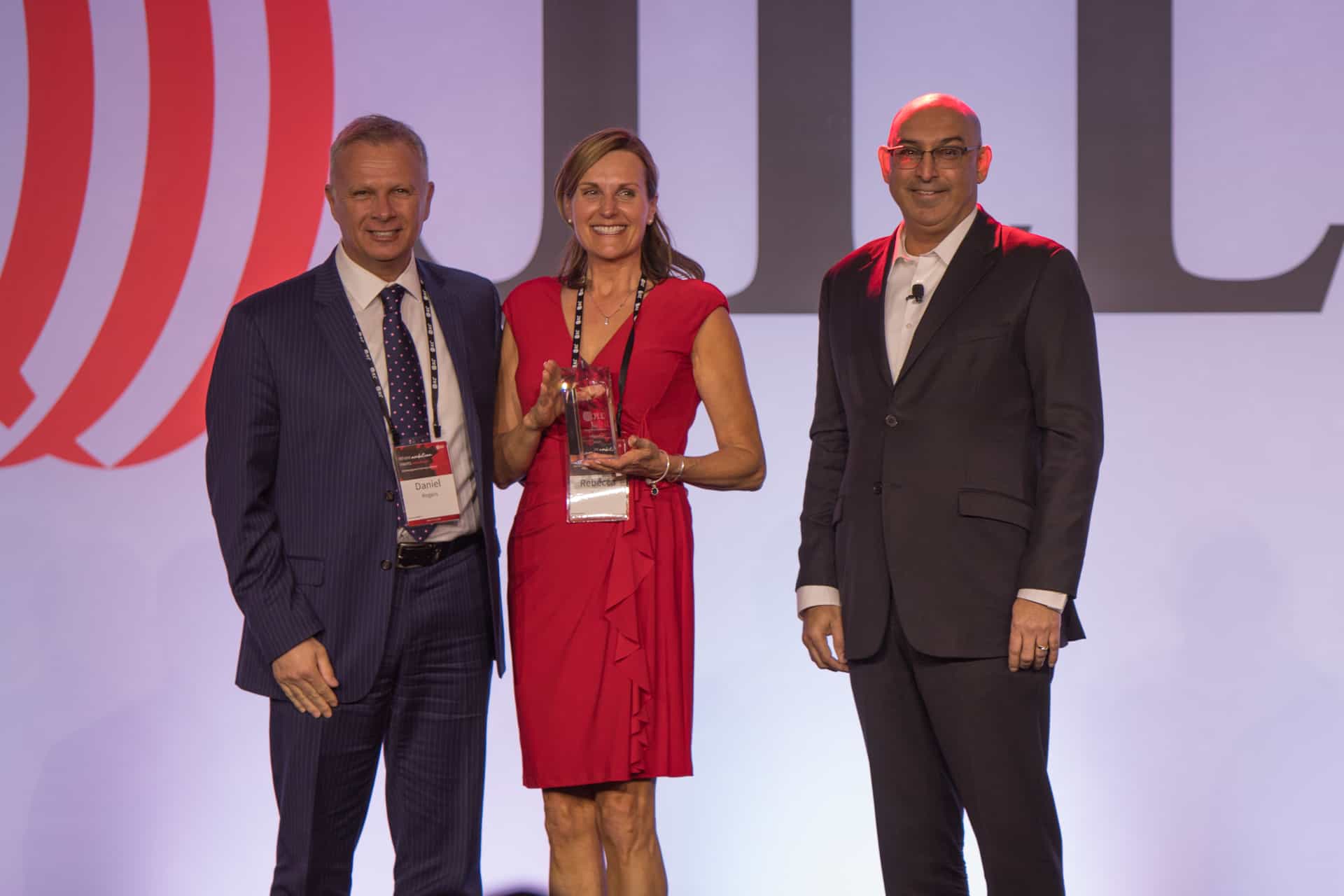 Three people stand smiling; the woman in a red dress holds an award. Men in suits flank her, with a partial logo on the backdrop.