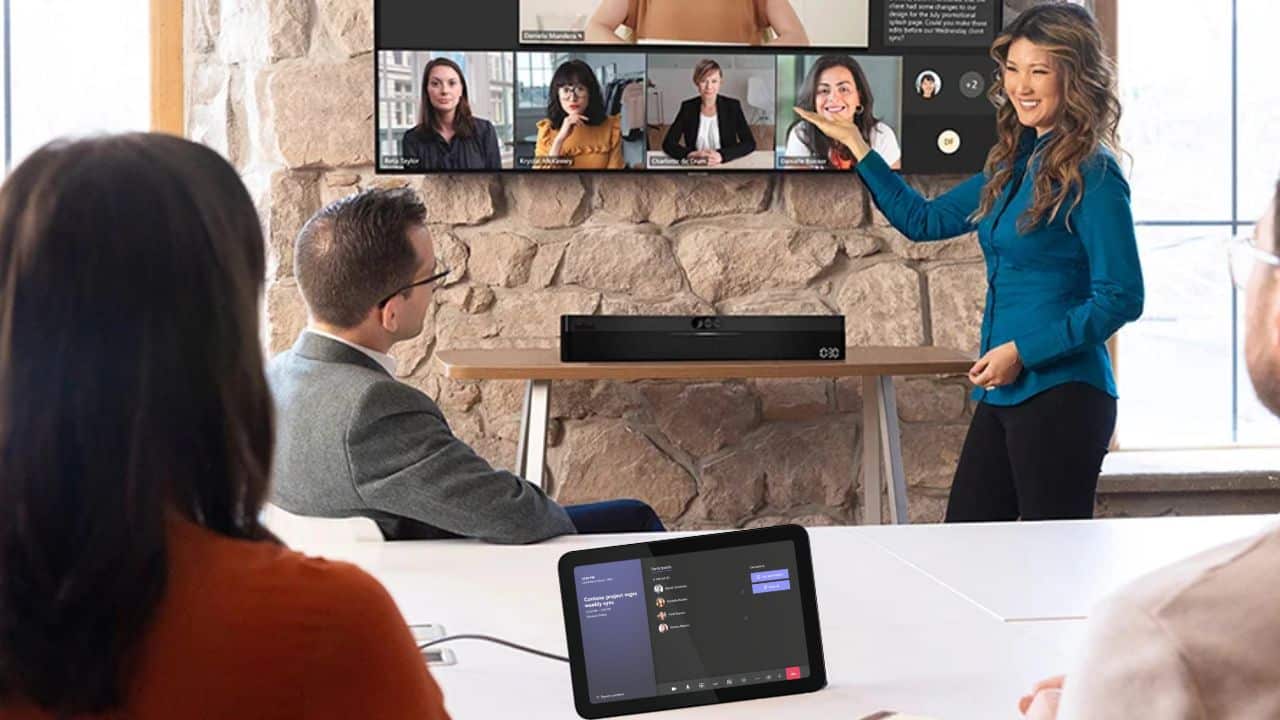 A woman in a blue blouse presents in a conference room with a video call screen, three listeners, and a tablet showing the interface.