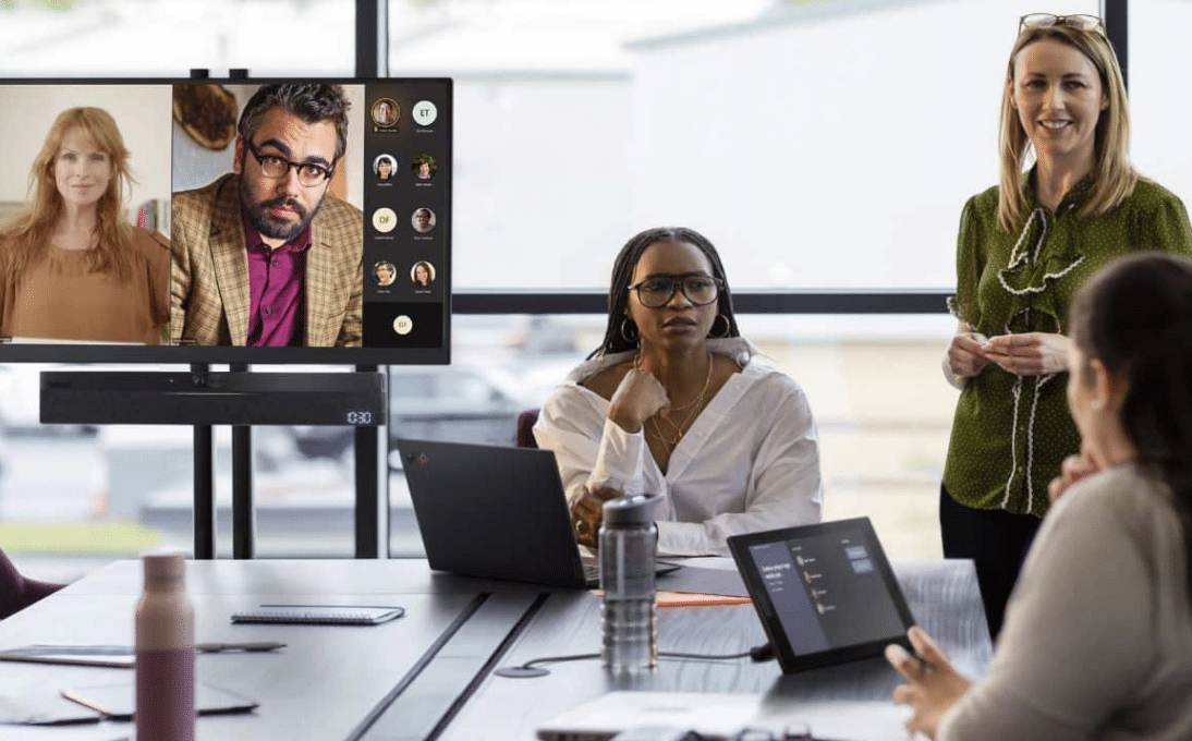 In a video conference, two participants on screen; four at the table with laptops. A woman speaks, windows providing natural light.