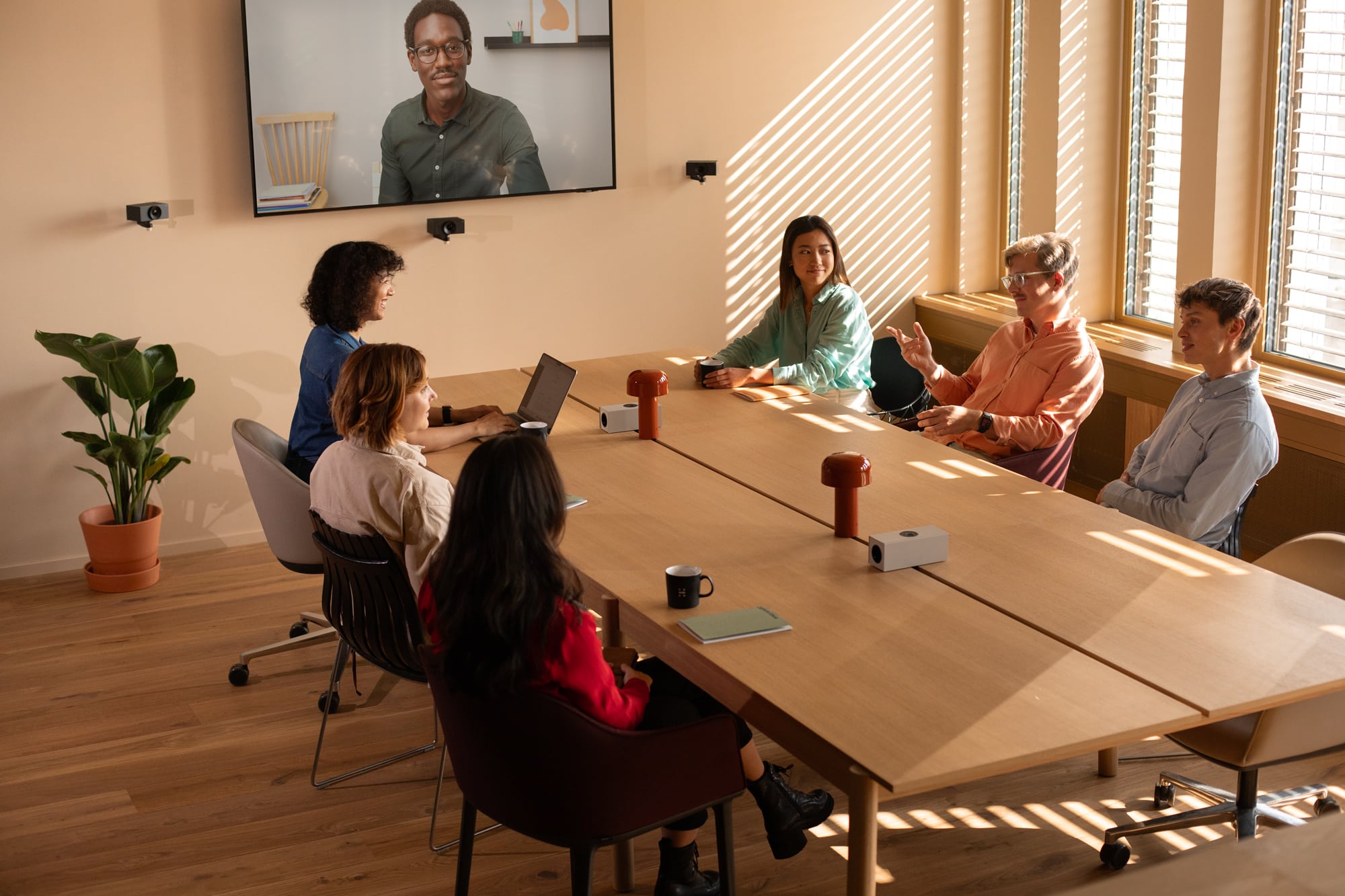 Six in a meeting: five at a table, one on screen. Laptop, mugs present. Sunlight and shadows; potted plant in corner.
