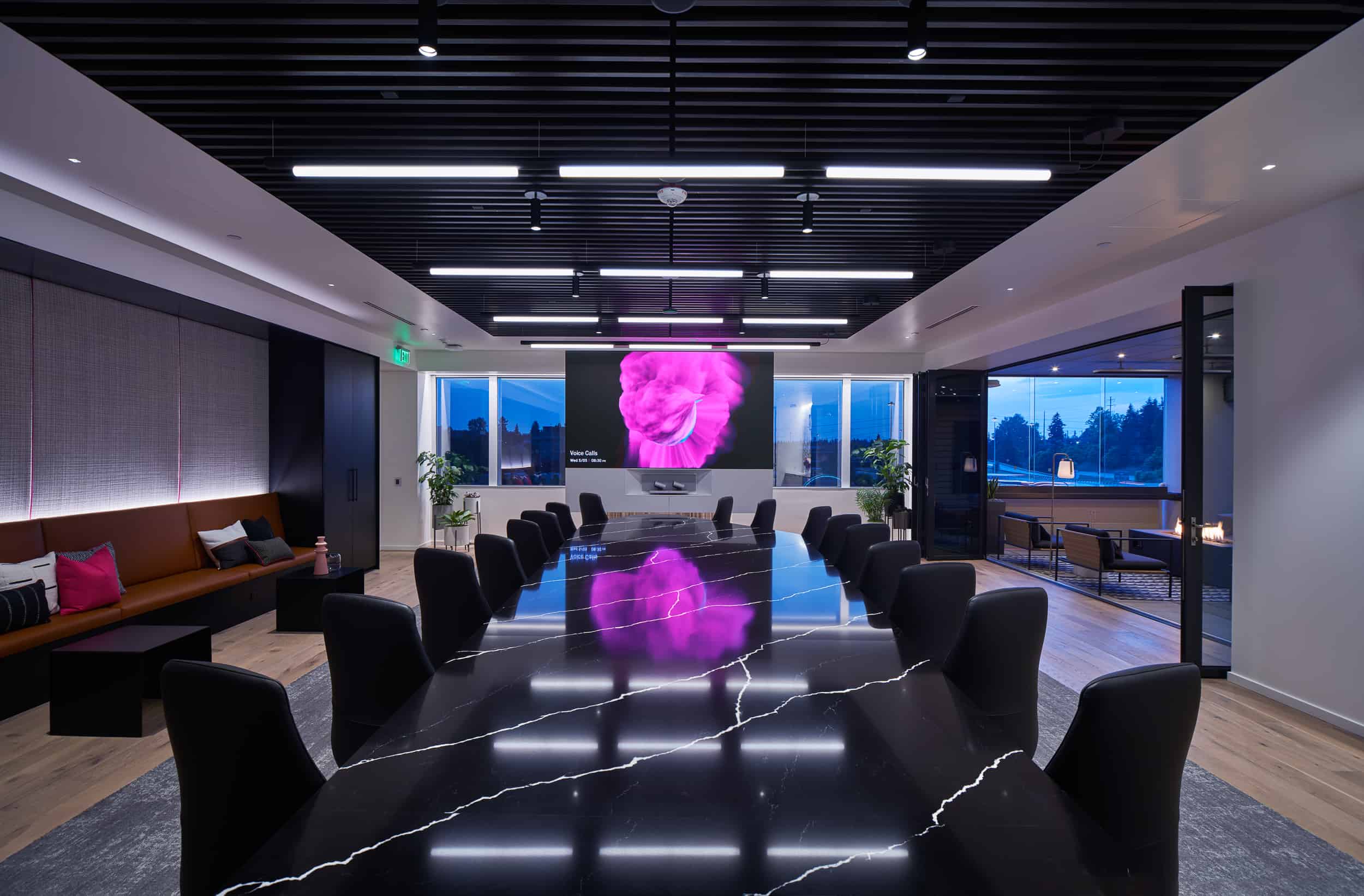 Modern conference room: glossy black table, black chairs, pink flower on screen, wood floors, plants, large windows with outdoor view.