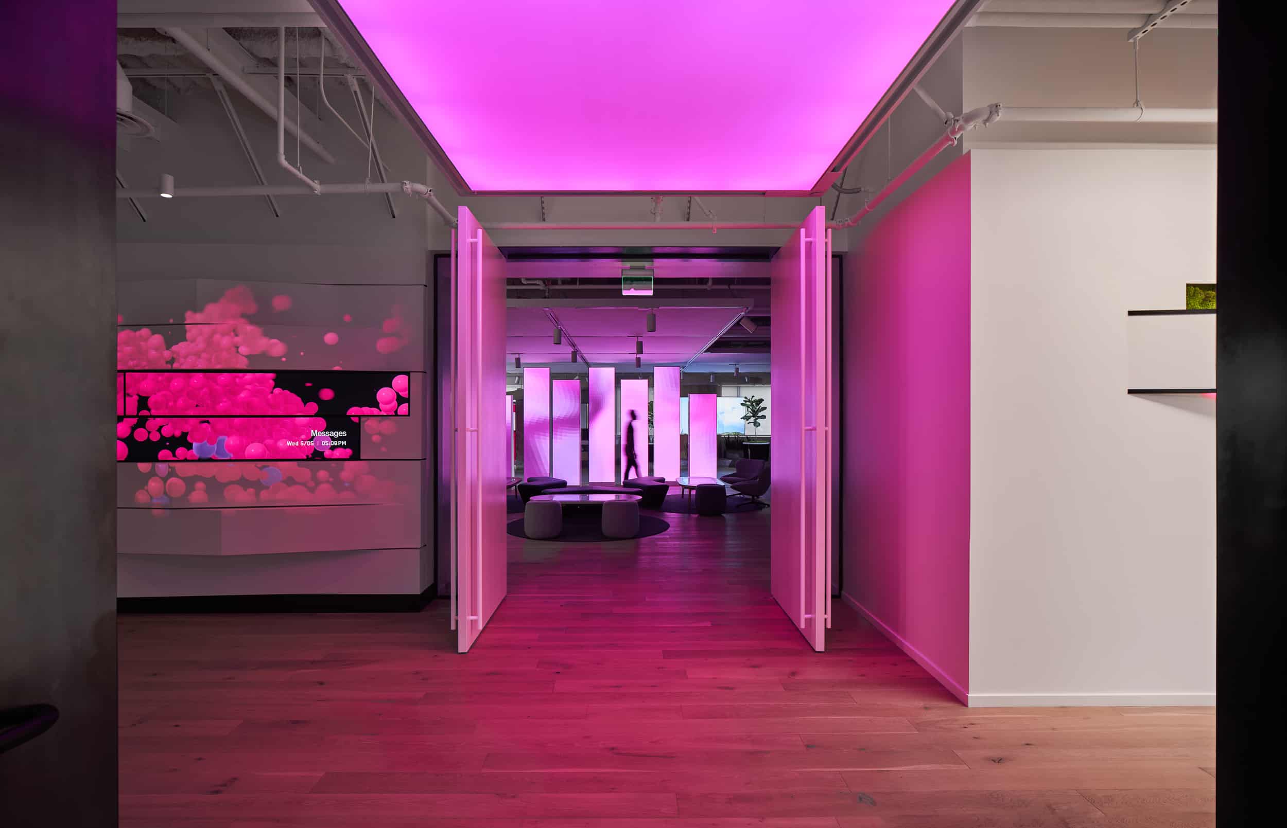Modern room: wooden floor, pink-lit ceiling, abstract shapes on screen; seating area with tables/chairs in ambient pink light.