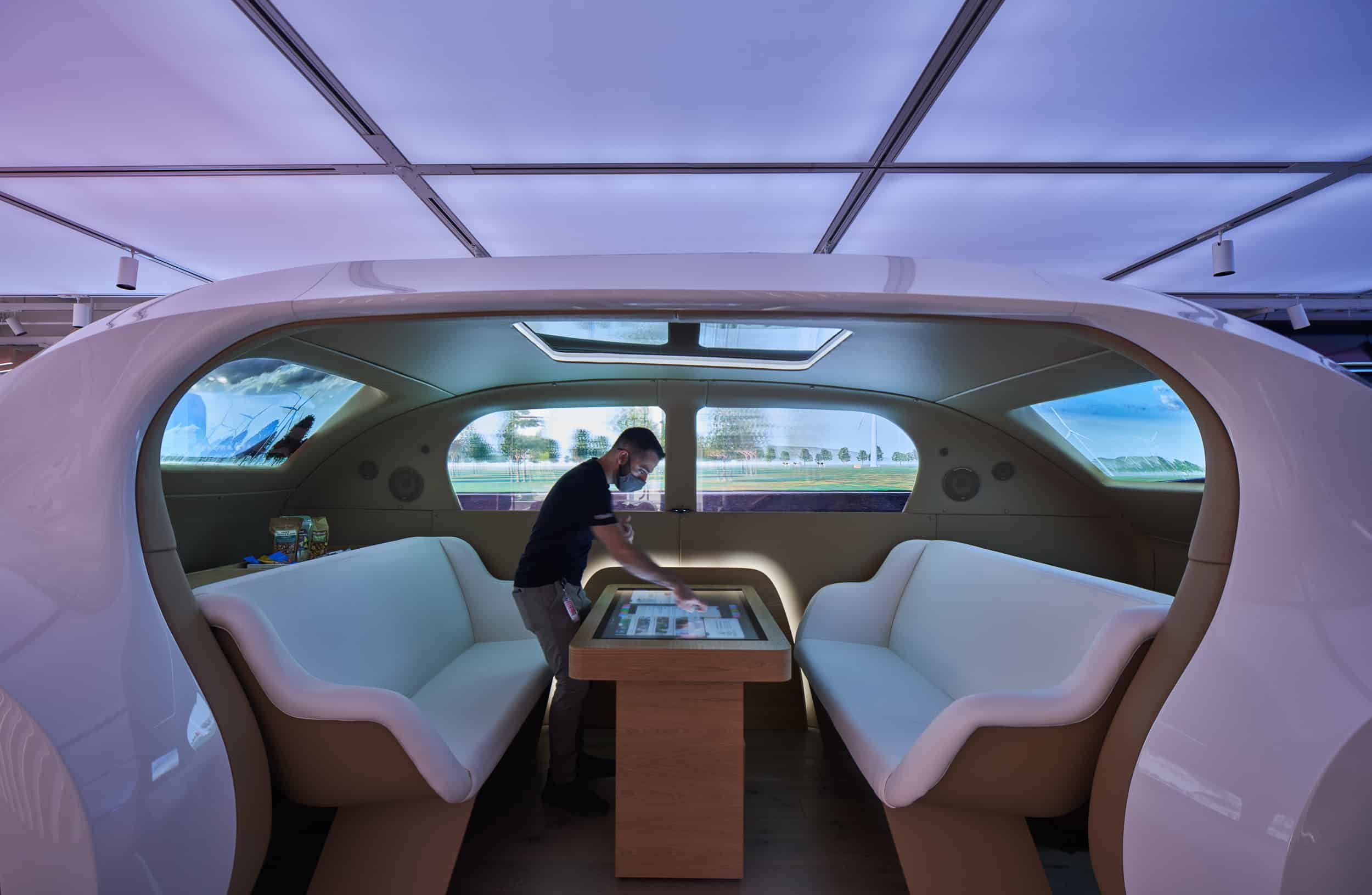 A sleek white interior with curved seats, central table. Person arranges items; windows show digital outdoor scenes.