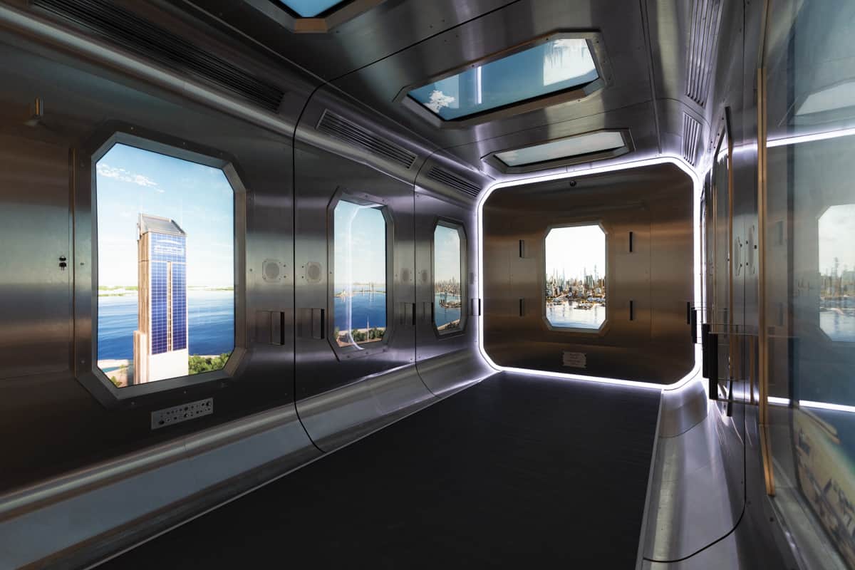 A sleek, metallic corridor with large windows revealing a cityscape of skyscrapers and water, accented by soft lighting and ceiling panels.