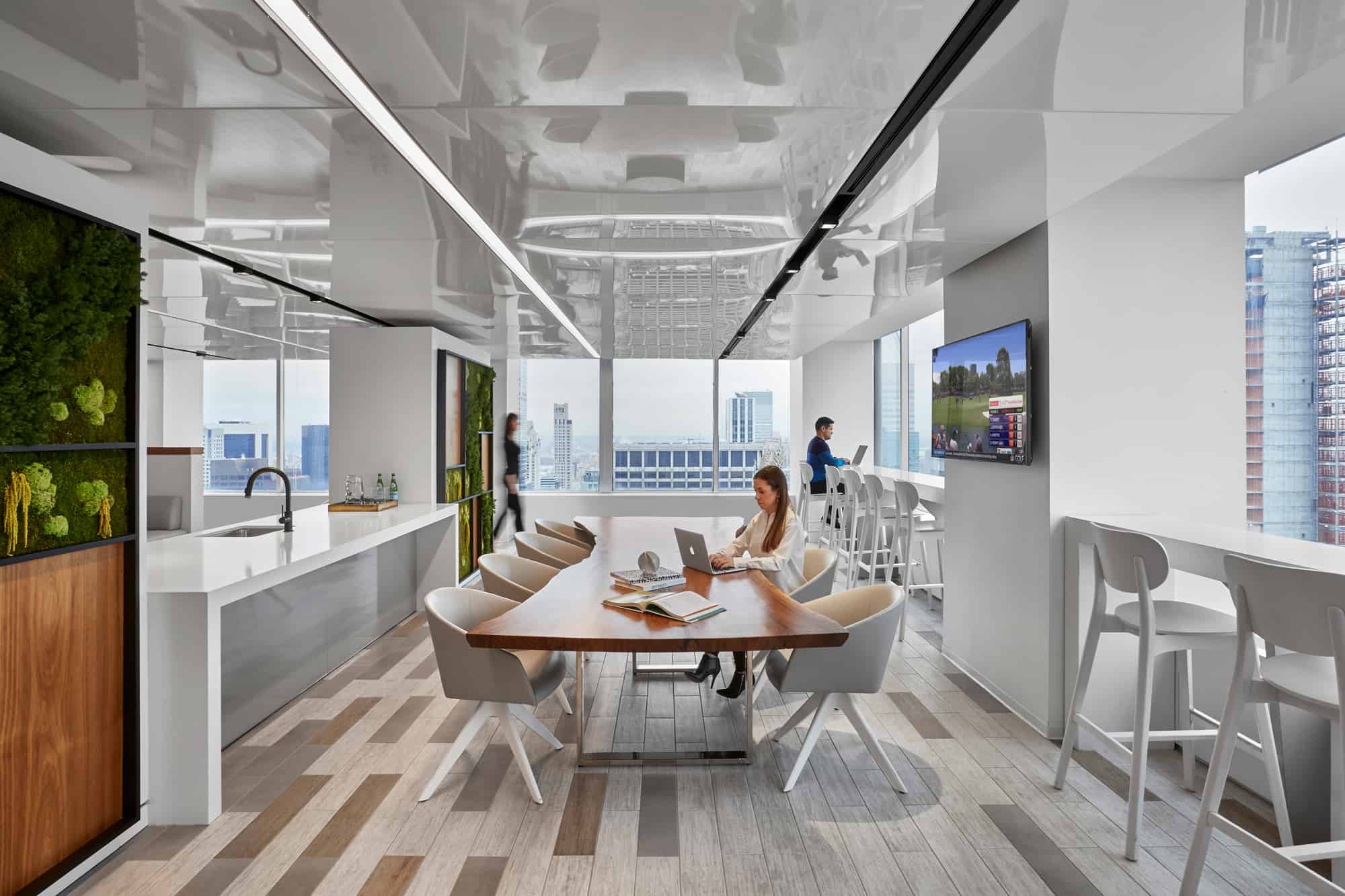 Modern office with a wooden table, laptop, city view, screen display, people interacting, and green plants.