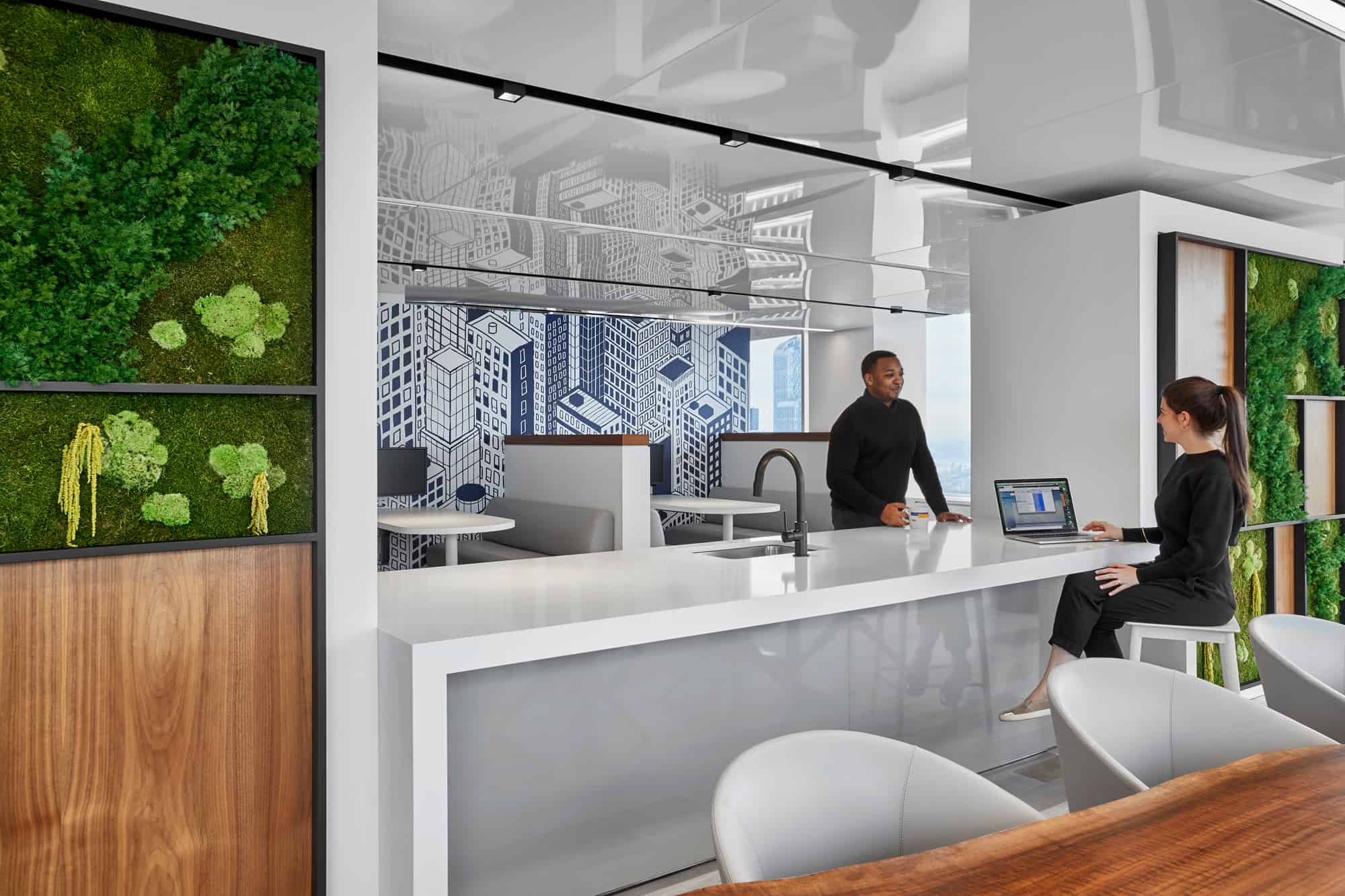 Modern office: two people interact by a white countertop with greenery, city art, and sleek furniture; one sits with a laptop.