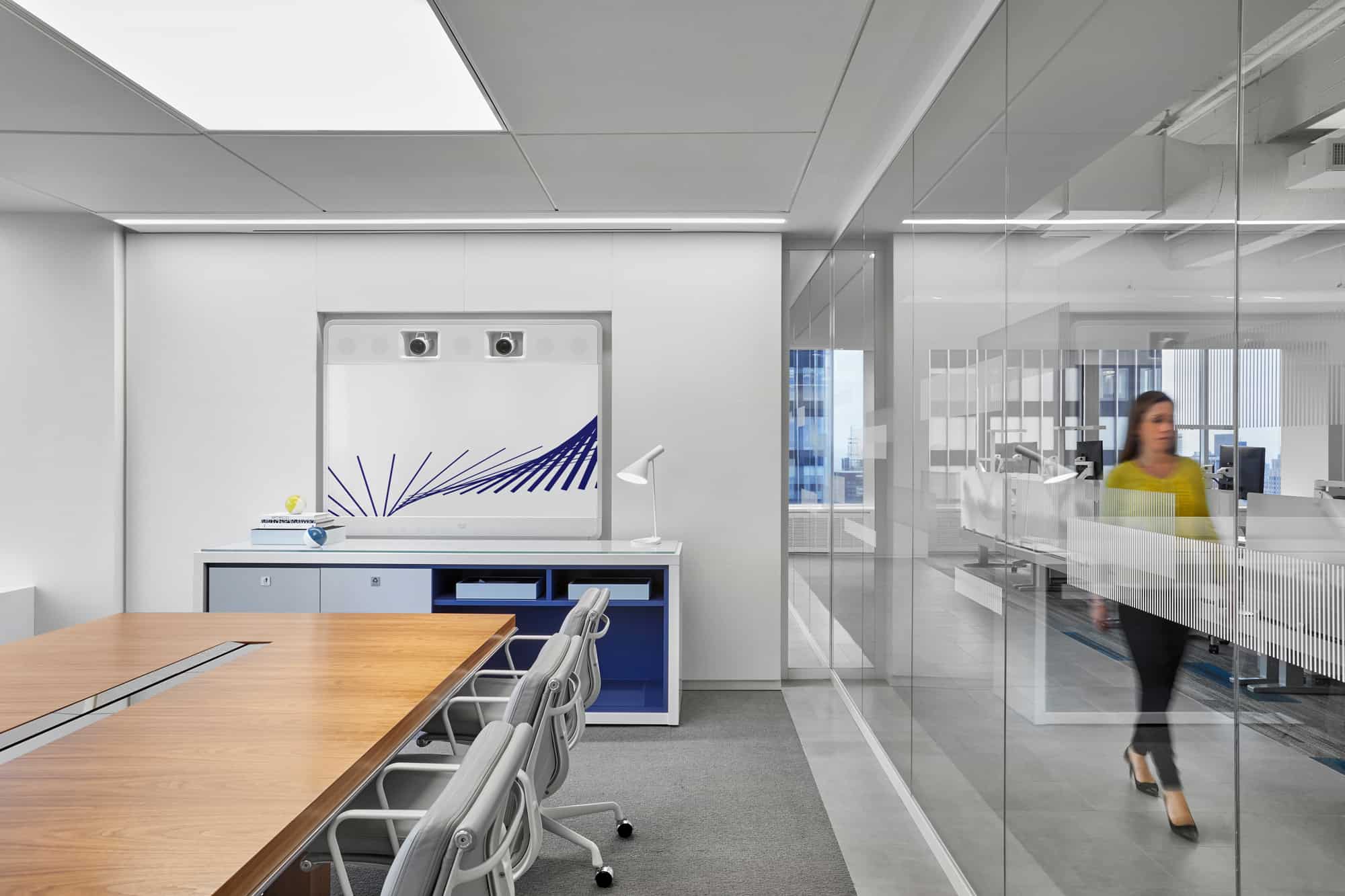 Modern office: conference table left, decorative cabinet with blue lines behind, woman in yellow top passes glass walls right.