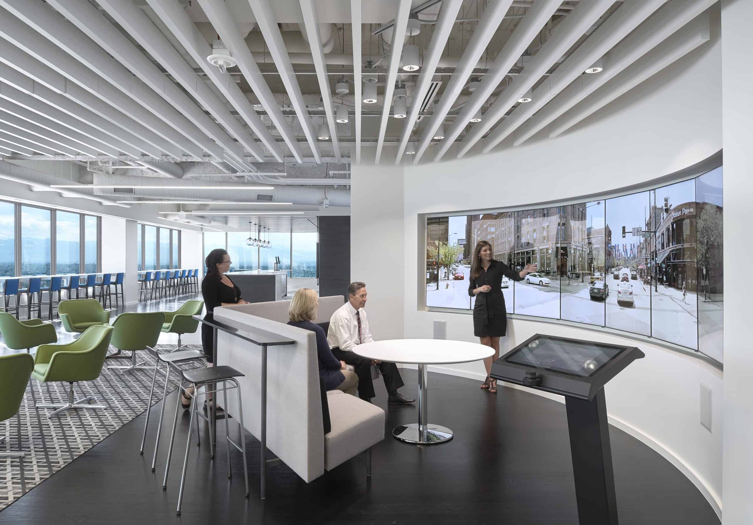 Modern office with 3 on curved sofa, 1 presenting at screens. Features: city displays, green chairs, tables, unique ceiling.