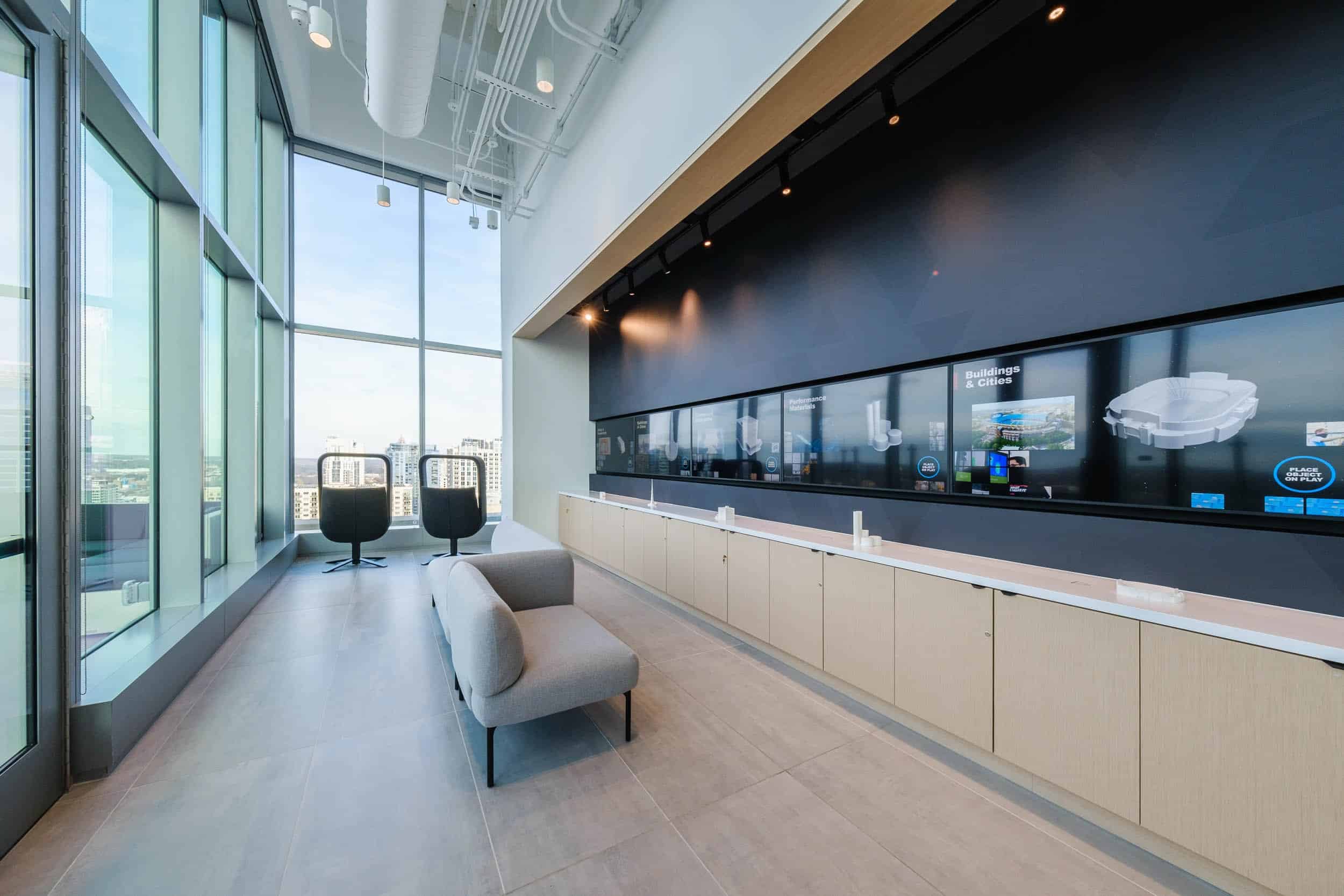 Modern office lounge: large windows, city view, sleek wood cabinetry, digital screen wall, gray armchair, two black chairs by windows.