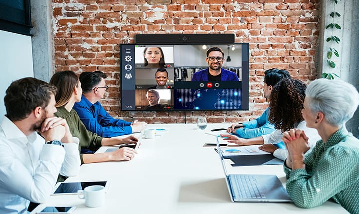 People in a conference room watching a video call with five participants; seated around a table with laptops, documents, and a plant.