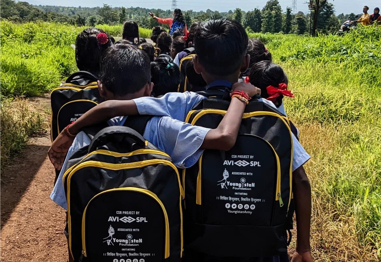 Children walk on a dirt path through green fields, in uniforms with black backpacks. One has an arm around another, under a blue sky.