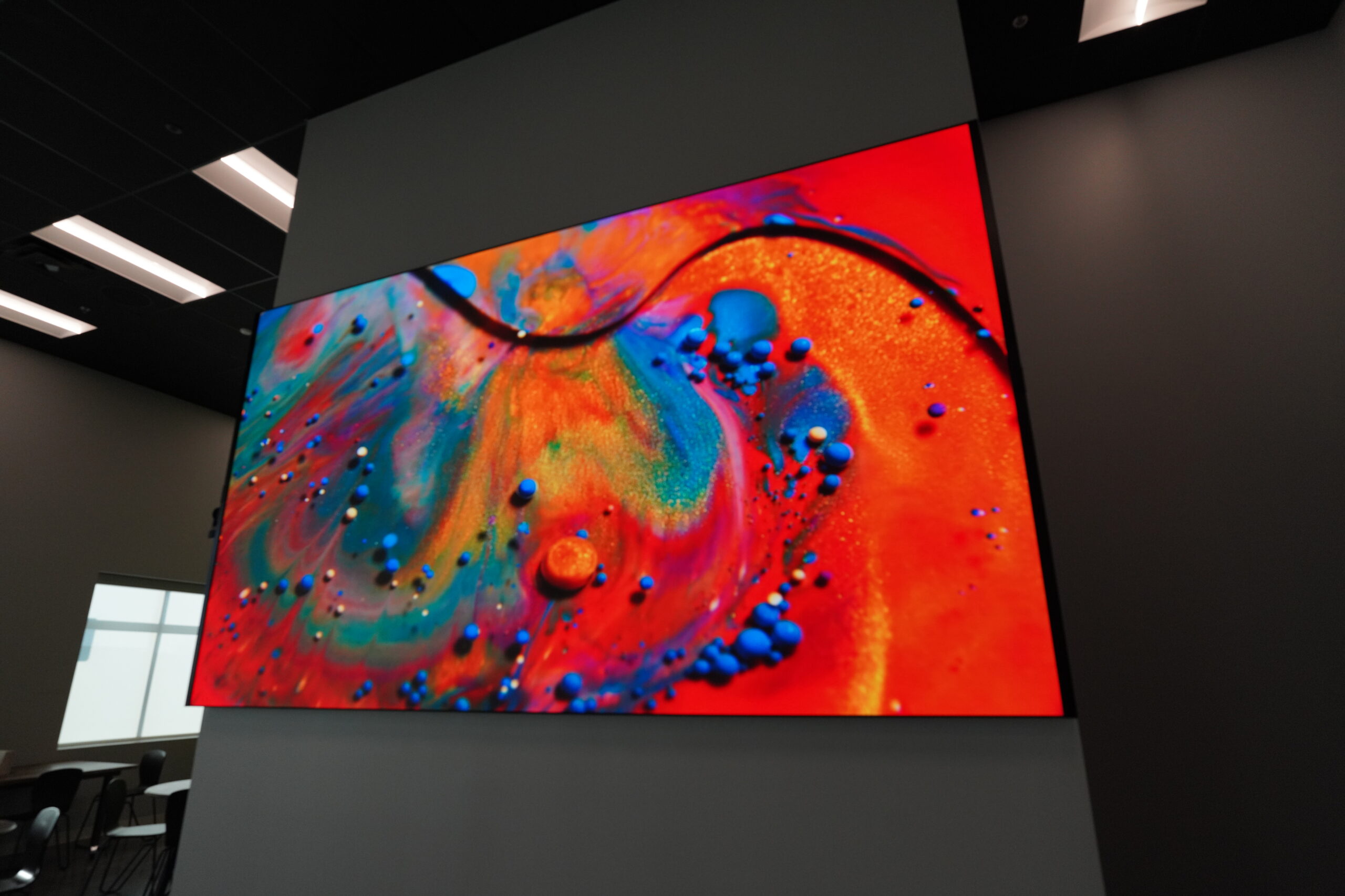 Wall-mounted screen shows vibrant abstract art in orange, blue, red; dimly lit background with ceiling lights and part of window visible.