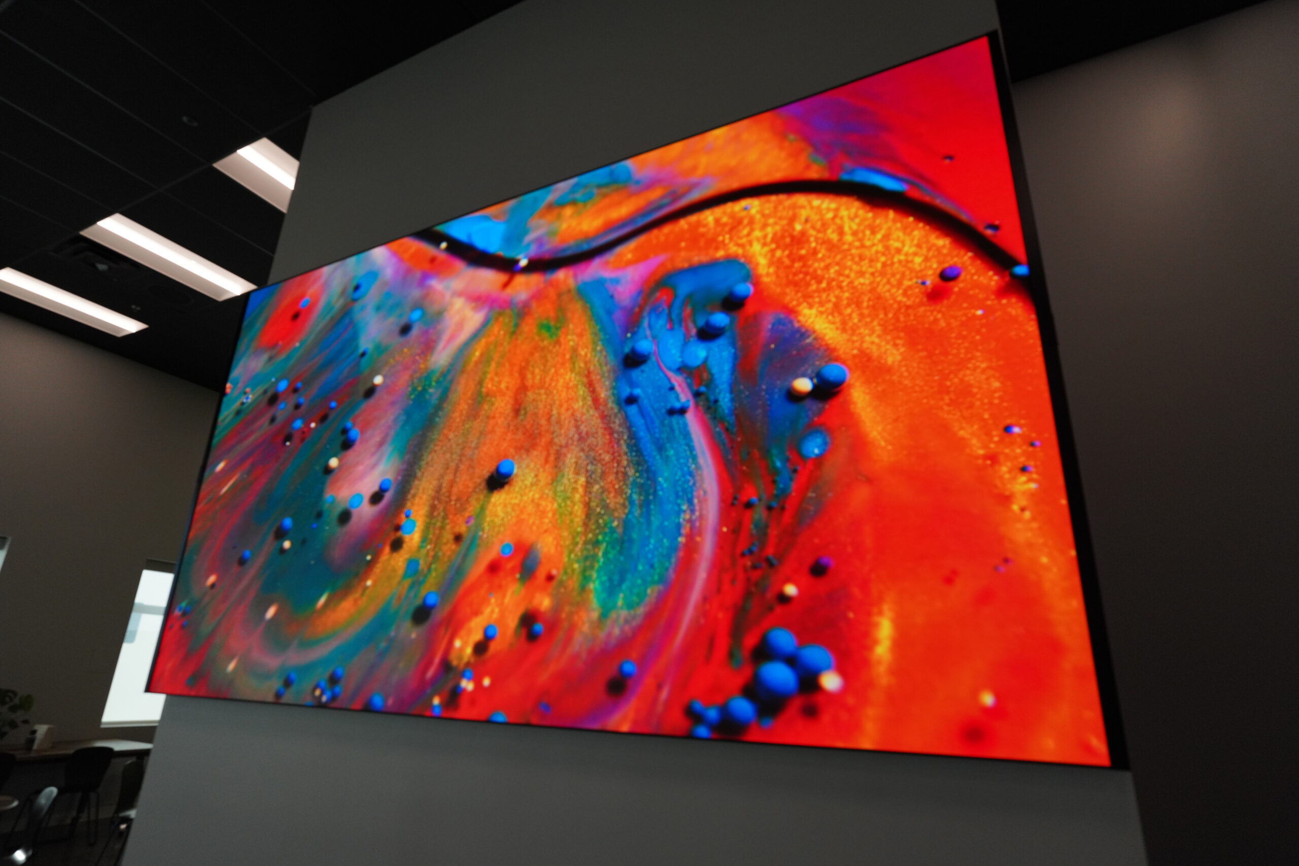 A large screen shows a vibrant abstract with swirls of red, orange, yellow, blue, green, and small blue dots under overhead lighting.
