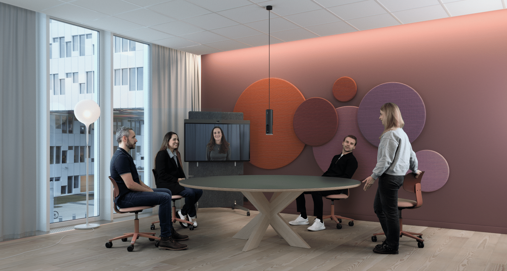 Four colleagues sit around a round table with a video call on screen. Pink walls, circular decor, large window with curtain.
