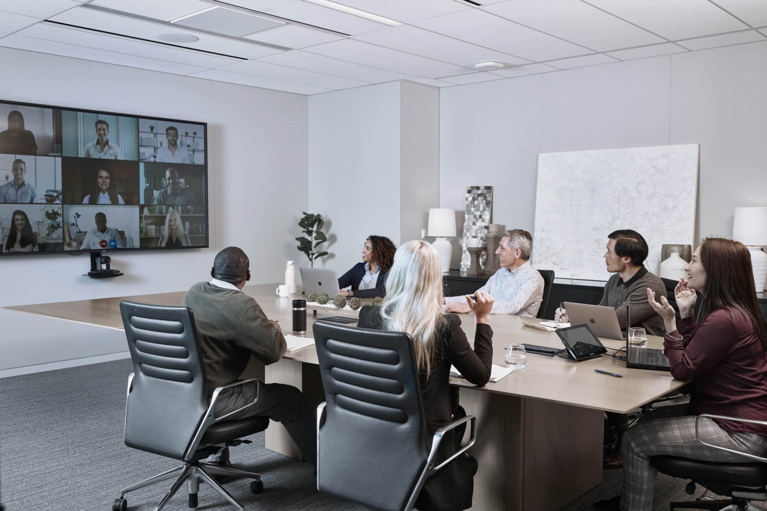 A group around a conference table engaged in a video meeting, interacting with the screen; modern decor with lamps and artwork.